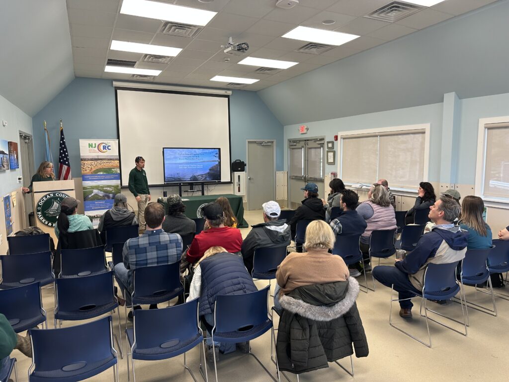 A group of about 20 audience members listens to a presenter standing in front of a slideshow