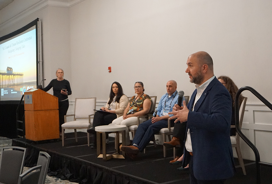 A standing speaker addresses the audience with seated panelists watching