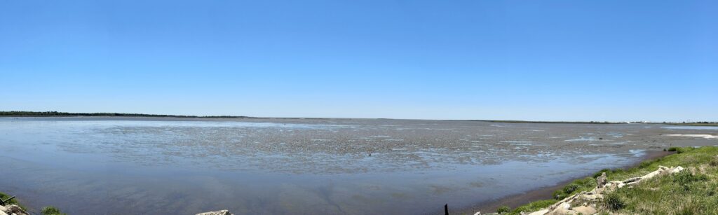 A panoramic view of a shallow, tidal waterfront, with wet mud and sand exposed in some areas. 